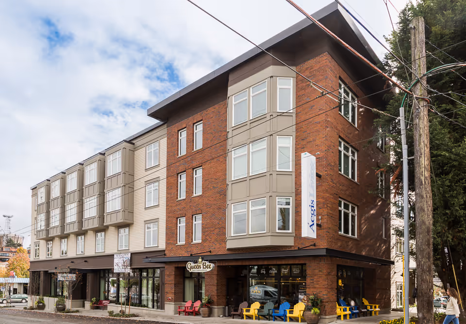 Exterior view of a multi-story brick and beige building with large windows and a sign that reads 'Aegis'. There are colorful chairs and potted plants outside the ground floor, which appears to house a business named 'Queen Bee'. A person is walking on the sidewalk near the building, and there are power lines and trees visible.