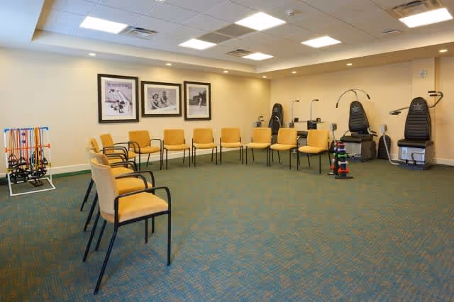 A spacious room with a circle of yellow cushioned chairs along the walls, exercise equipment including resistance bands, weights, and two seated exercise machines, and three black and white framed photos on the wall under bright ceiling lights.