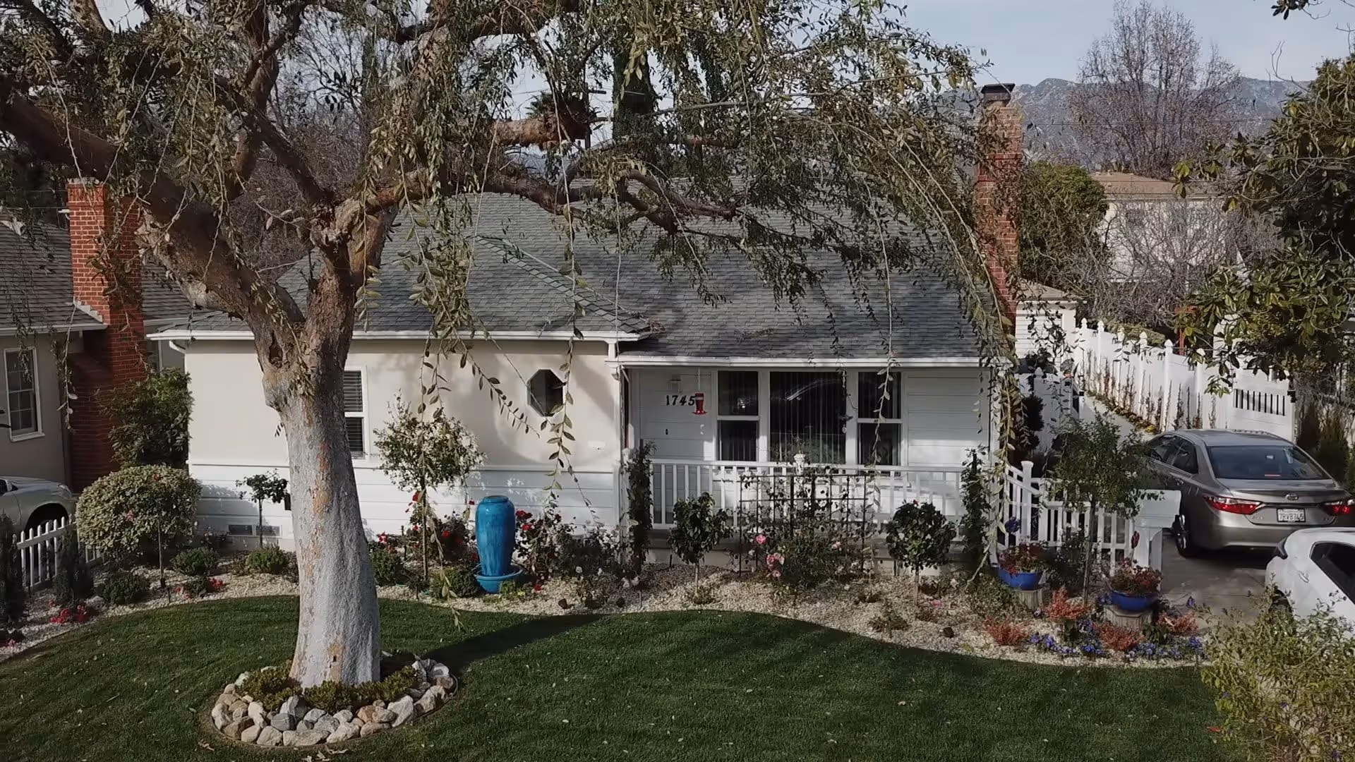 Front exterior view of a single-story house with a gray roof, white walls, and a small porch. The house number 1745 is visible near the entrance. There is a large tree with hanging branches in the front yard, surrounded by a well-maintained lawn and various plants and flowers. Two cars are parked on the driveway to the right of the house, and a white picket fence lines the property.