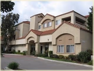 Exterior view of a multi-story residential building with beige stucco walls and red tile roofing, surrounded by small shrubs and trees under a partly cloudy sky.