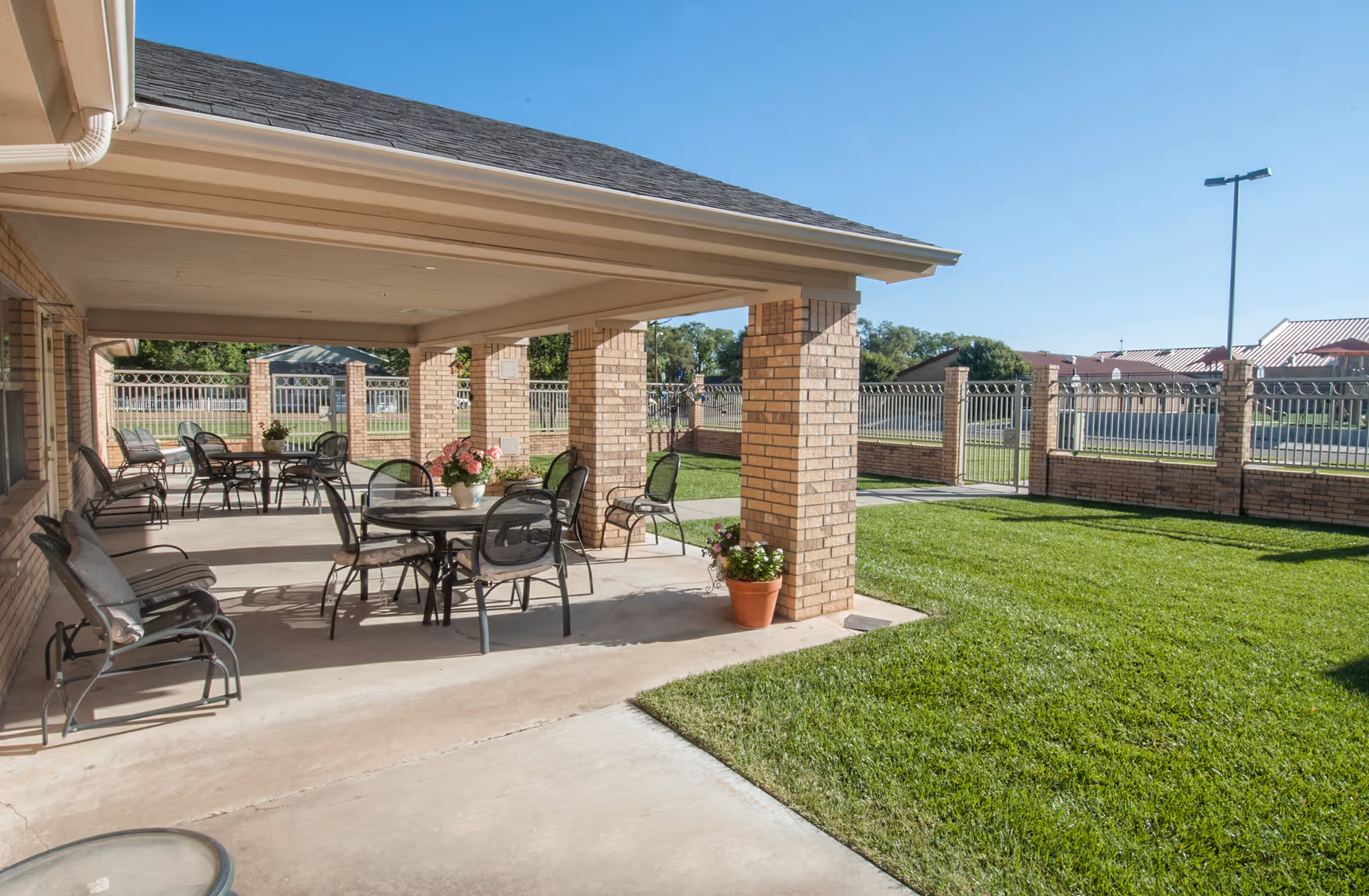 Covered outdoor patio area with multiple tables and chairs, potted plants on the ground and tables, adjacent to a grassy lawn enclosed by a brick and metal fence under a clear blue sky.