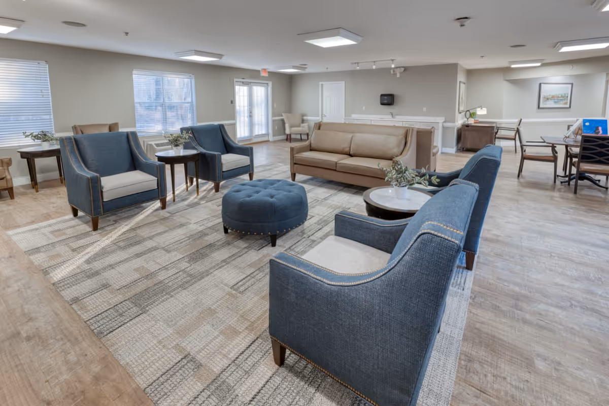 Bright, spacious lounge with blue armchairs, a tan sofa, a round ottoman and side tables on a patterned rug.