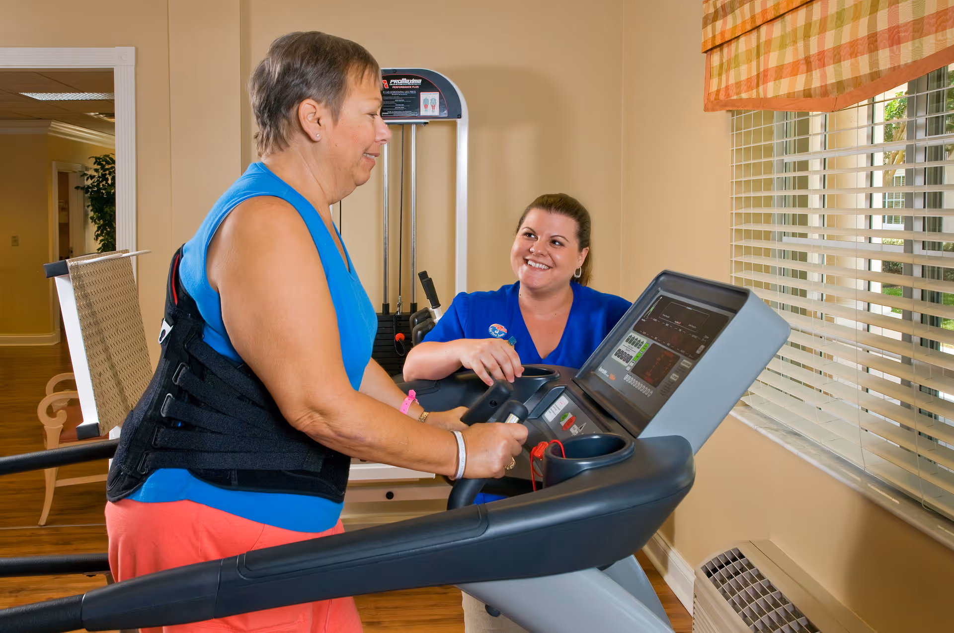 An elderly woman wearing a back support brace is walking on a treadmill while a smiling female staff member in blue scrubs watches and encourages her in a well-lit exercise room with a window and exercise equipment.