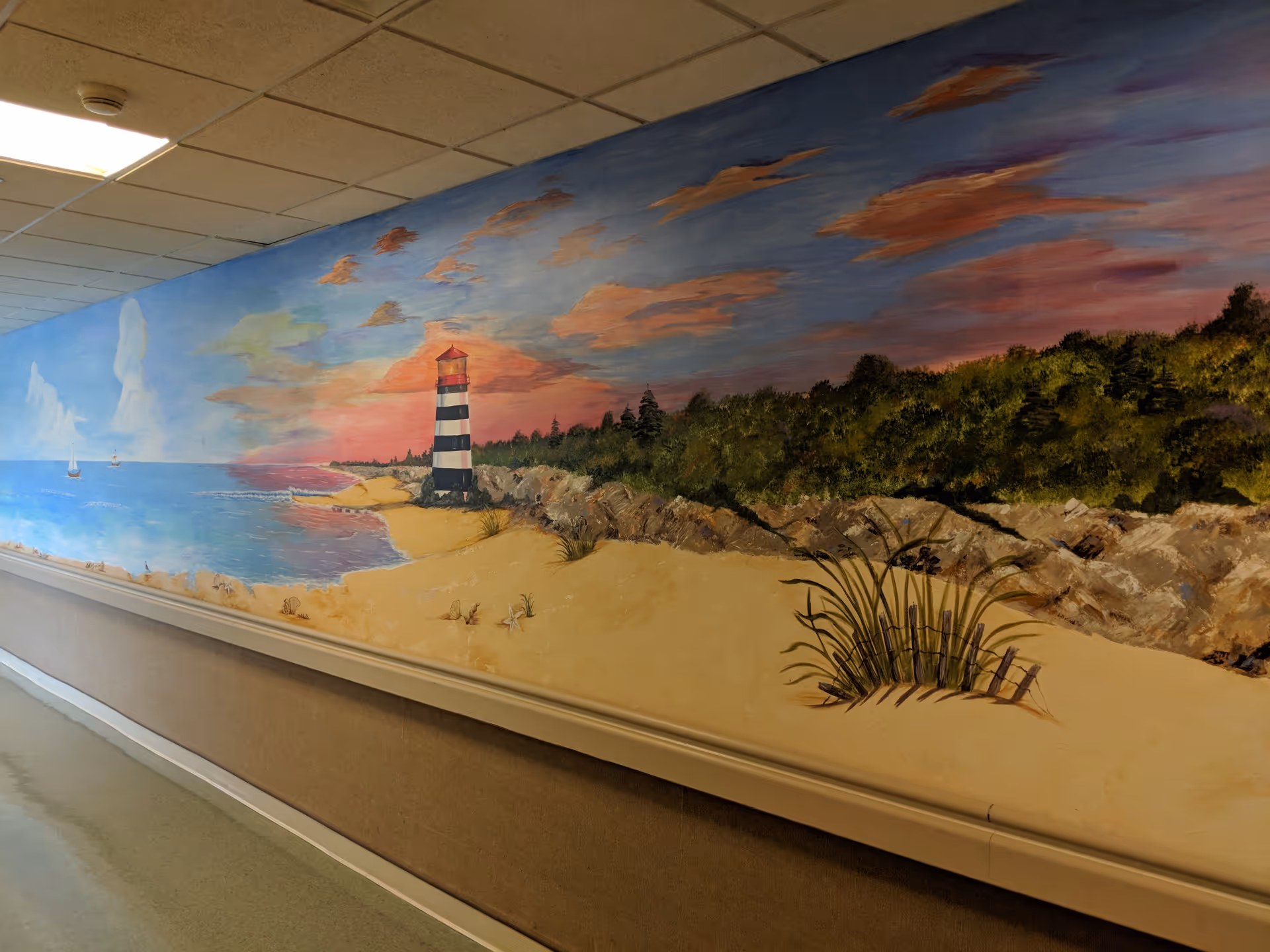 A painted mural on an interior wall depicting a beach scene with sand dunes, grass, a lighthouse, ocean waves, sailboats, and a colorful sky at sunset or sunrise.
