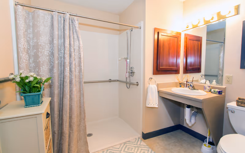 A clean and well-lit bathroom featuring a walk-in shower with a floral-patterned shower curtain, a beige countertop with a sink, a mirror with wooden cabinets above, a white towel hanging on a ring, a toilet, and a small cabinet with a green potted plant on top.