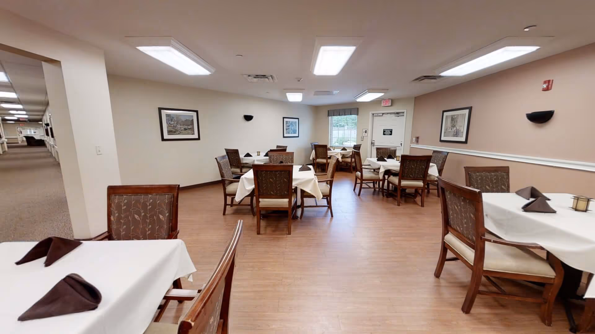 A dining room with several square tables covered with white tablecloths and brown folded napkins. Each table is surrounded by wooden chairs with patterned cushions. The room has light-colored walls with framed pictures, wood flooring, and bright ceiling lights. A window and an exit door are visible at the far end of the room.