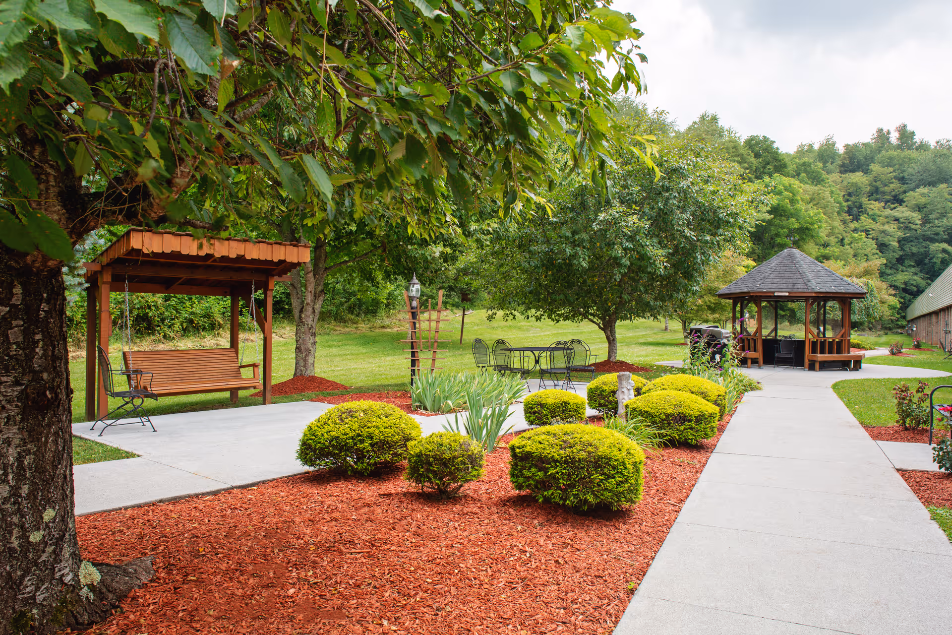 Landscaped outdoor courtyard with a paved walkway, gazebo, wooden swing, metal patio table, and trimmed shrubs.