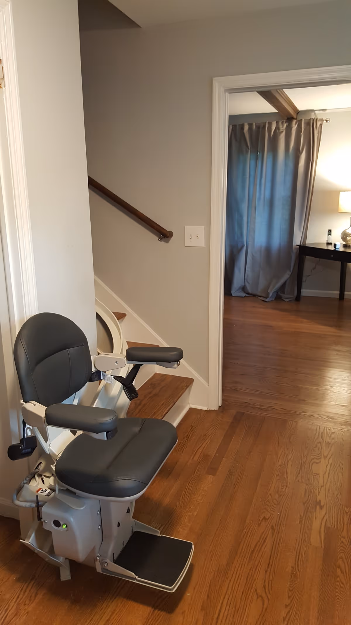 A stairlift chair installed on a wooden staircase inside a home. The chair is gray with armrests and a footrest. The room has wooden flooring and light-colored walls. In the background, there is an open doorway leading to another room with a window covered by gray curtains and a table with a lamp on it.