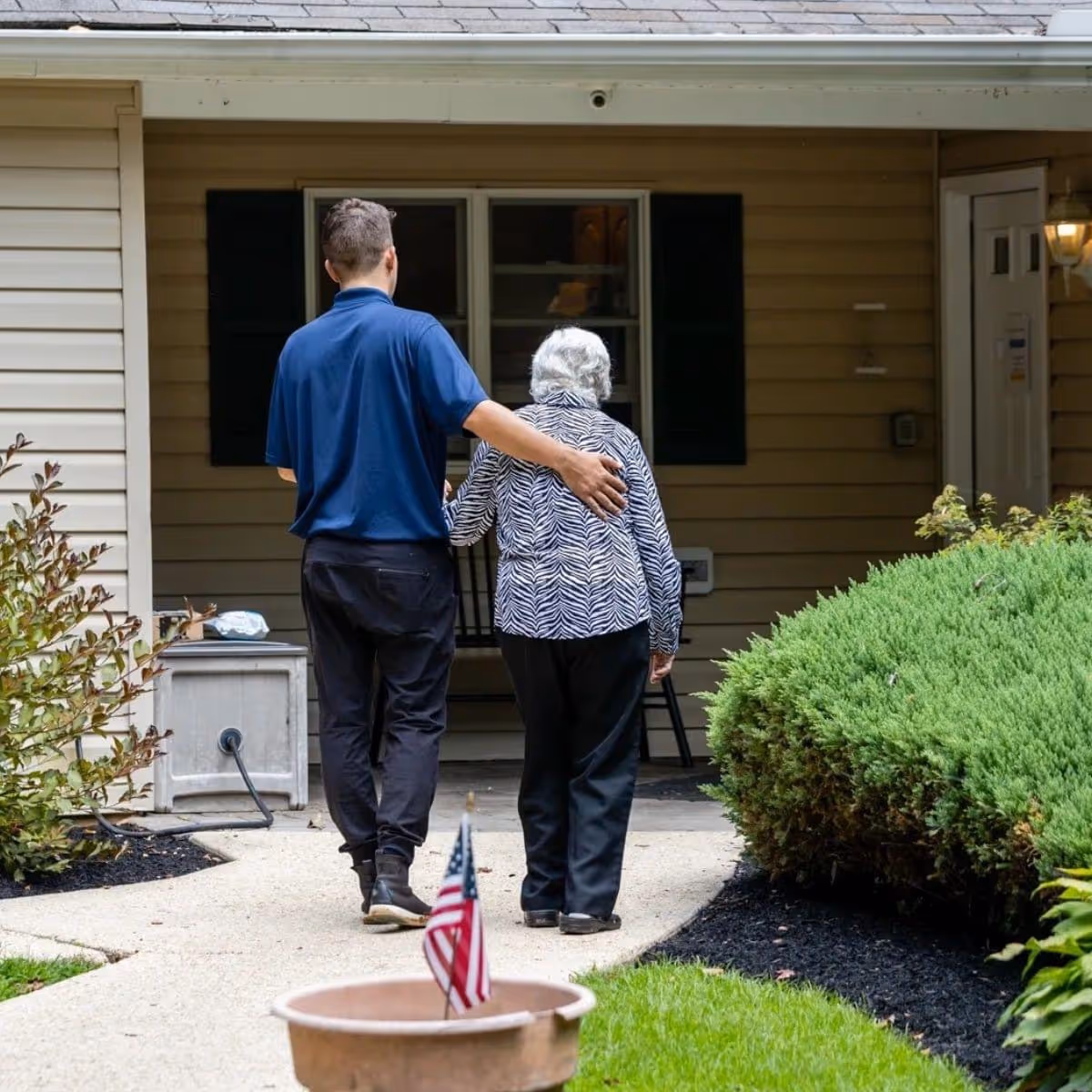 A caregiver in a blue shirt is walking arm-in-arm with an elderly woman wearing a zebra-striped jacket along a paved pathway towards the entrance of a beige building with black shutters. There are green bushes and a small American flag in a pot near the pathway.