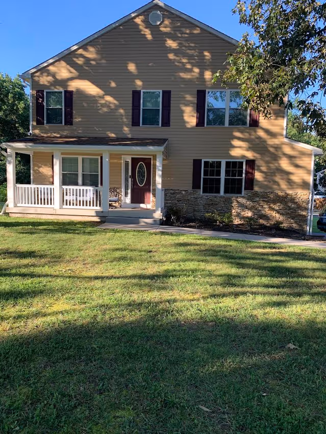 Two-story beige house with a front porch, maroon door, and a grassy lawn in front.