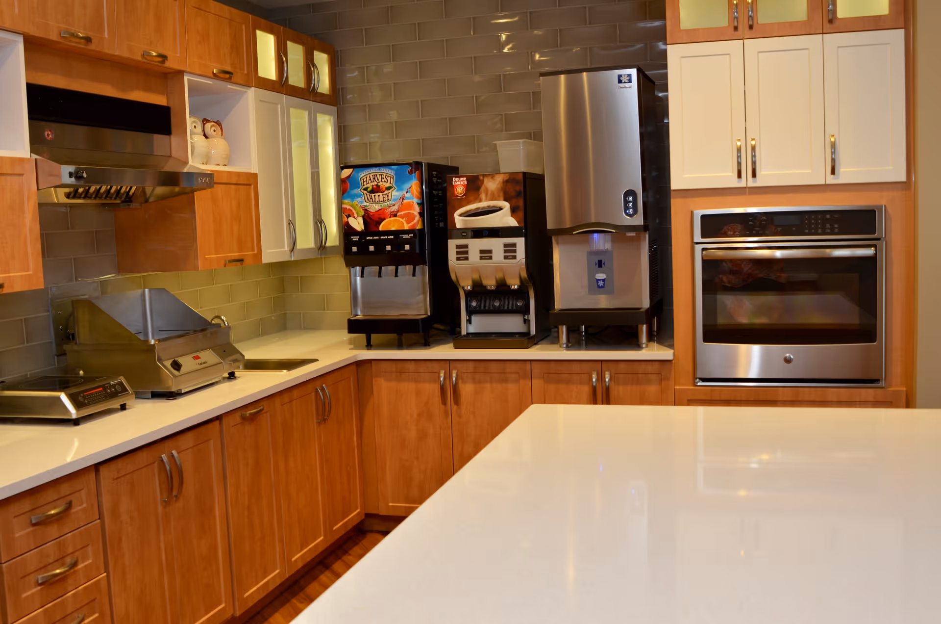 A modern kitchen area with wooden cabinets and white countertops. The kitchen features a stainless steel oven, a beverage dispenser with coffee and juice options, a water dispenser, and a small sink. The backsplash is made of glossy beige tiles.