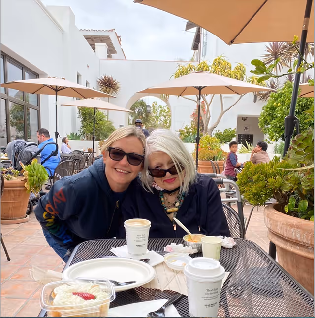 Two women wearing sunglasses sitting closely together at an outdoor table with coffee cups and dessert plates. They are smiling and appear to be enjoying a sunny day in a courtyard with large potted plants and umbrellas providing shade. Other people, including a child, are visible in the background.
