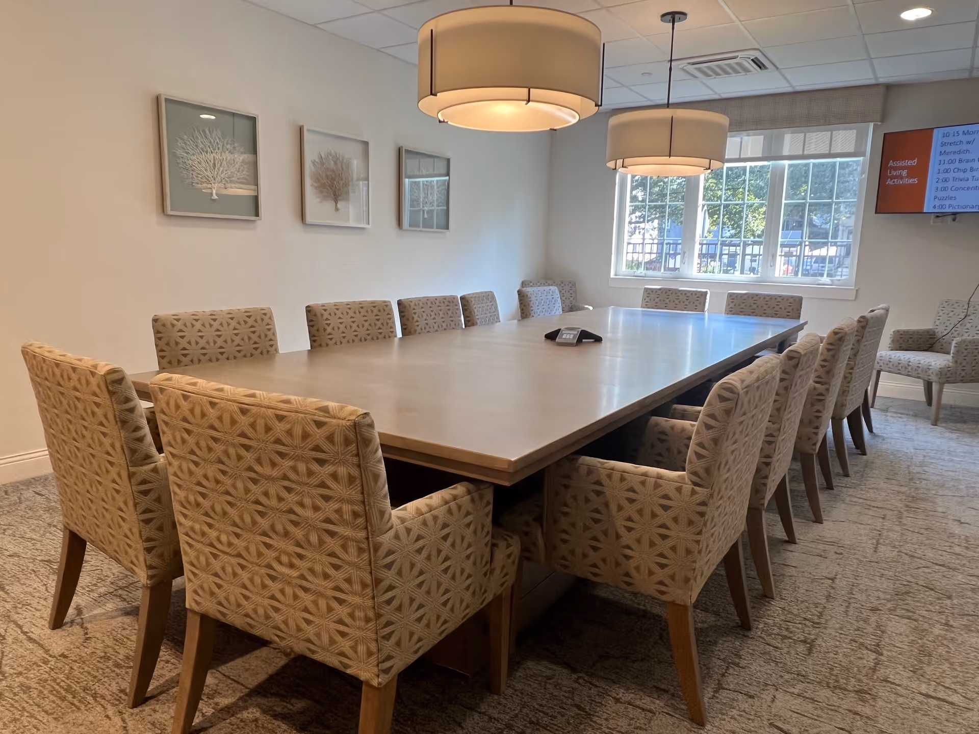 A conference room with a large rectangular table surrounded by twelve patterned upholstered chairs. Two large pendant lights hang above the table. The room has a window letting in natural light, three framed coral artwork pieces on the wall, and a wall-mounted TV screen displaying a schedule.