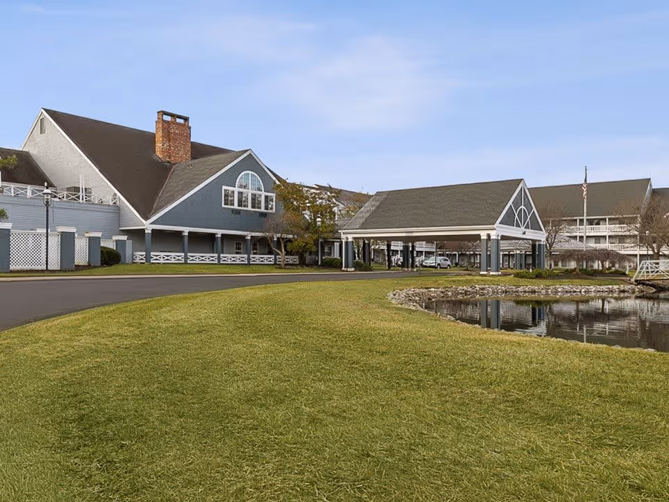 Exterior view of a senior living facility with blue and white buildings, a covered entrance, a small pond with a stone border, and a well-maintained grassy area under a clear sky.