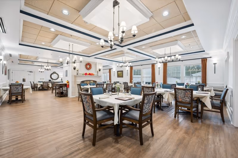 A spacious and well-lit dining room with multiple tables covered in white tablecloths, each surrounded by wooden chairs with patterned cushions. The room features large windows with brown curtains, chandeliers hanging from a coffered ceiling, and a fireplace decorated with a wreath and seasonal ornaments.