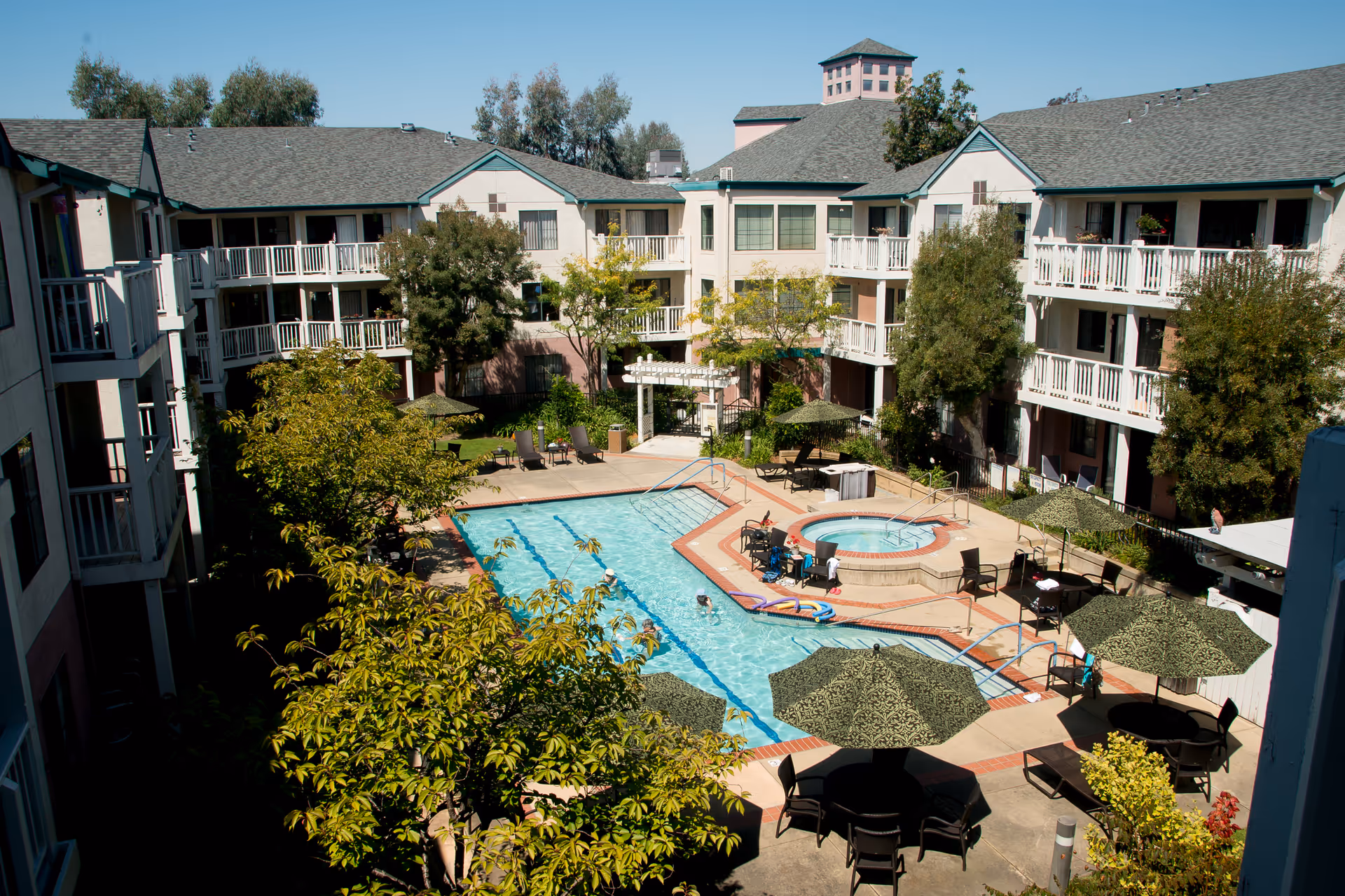 A three-story senior living building surrounding a central courtyard with a swimming pool, hot tub, umbrellas and seating.