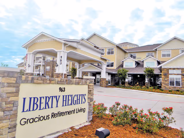 Exterior view of Liberty Heights Gracious Retirement Living facility showing the main entrance with a covered driveway, a large sign with the facility name and address, and a landscaped area with flowers and shrubs in front.