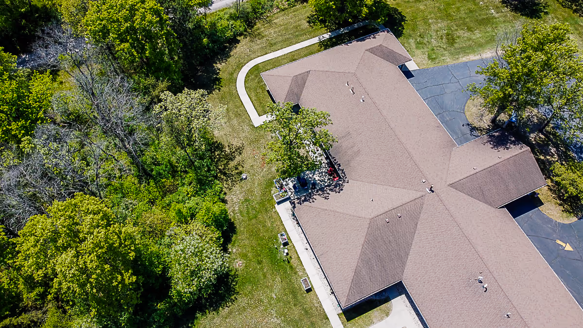 Aerial view of a single-story building with a brown pitched roof, adjacent parking, walkways and surrounding trees and lawn.