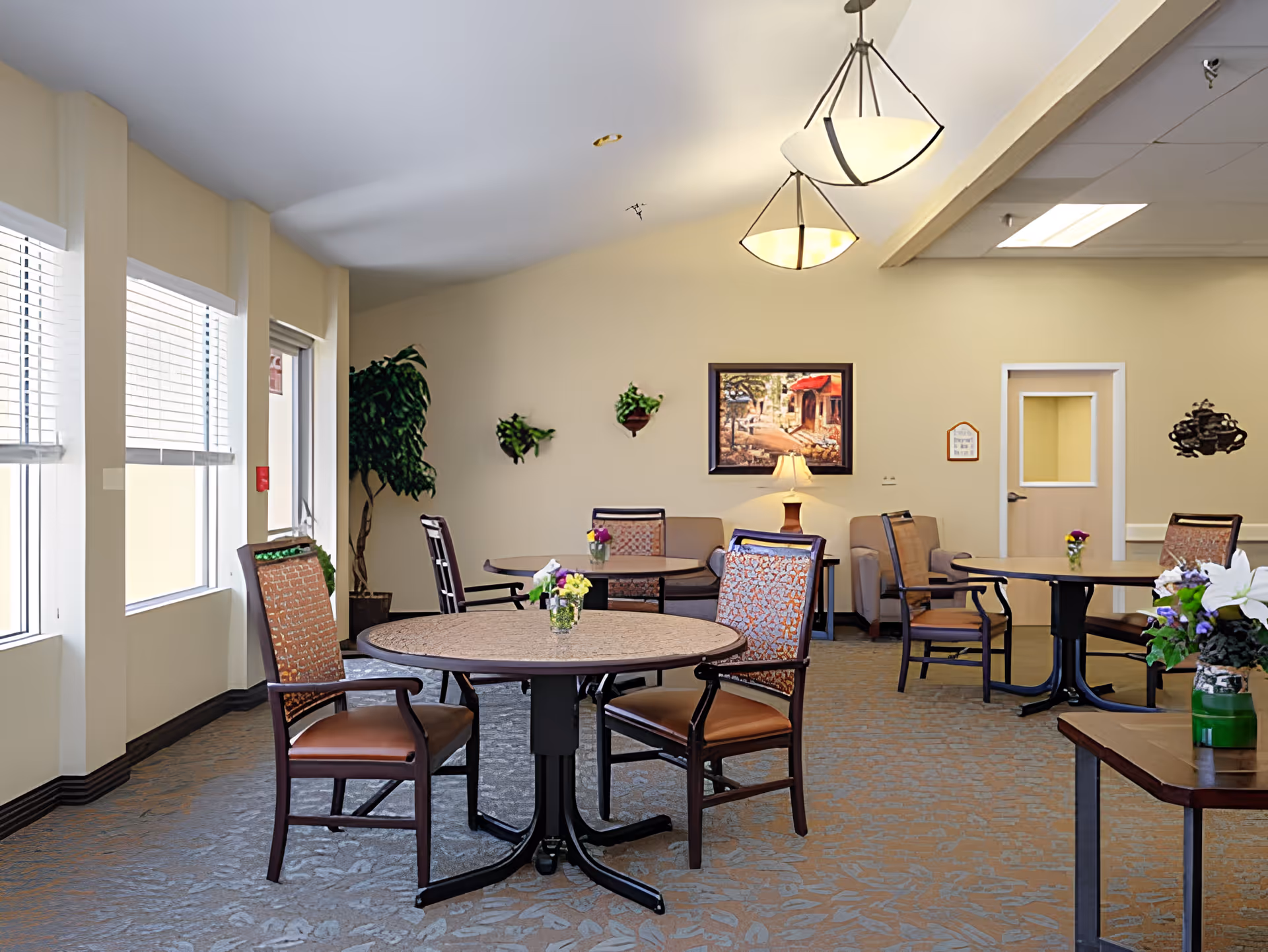 Bright communal dining room with round tables and chairs, potted plants and wall art.