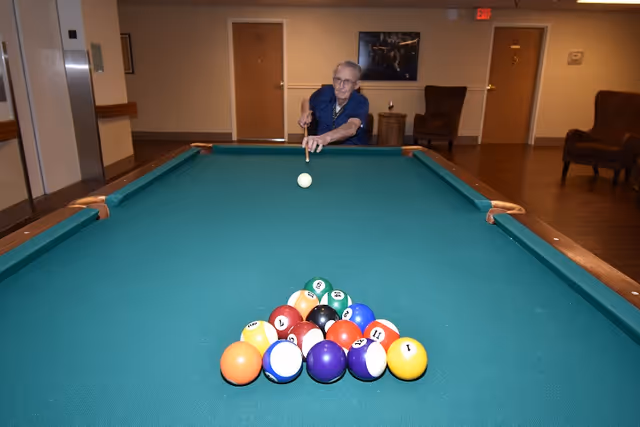 An elderly person playing pool in a common area with a green pool table, wooden floor, and chairs in the background.
