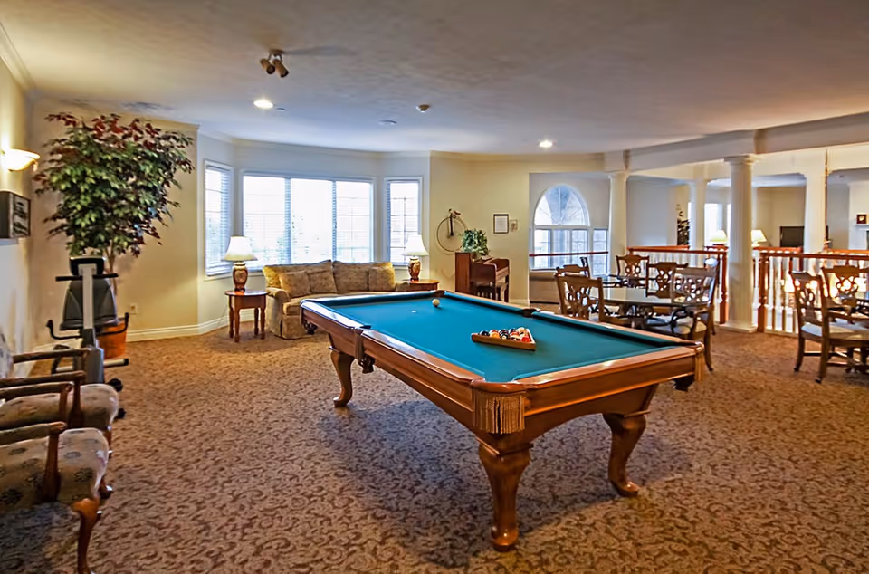 A spacious common area with a pool table in the center, surrounded by chairs and tables. There is a sofa and side tables with lamps near large windows letting in natural light. The room has carpeted floors, decorative columns, and a piano against the wall.