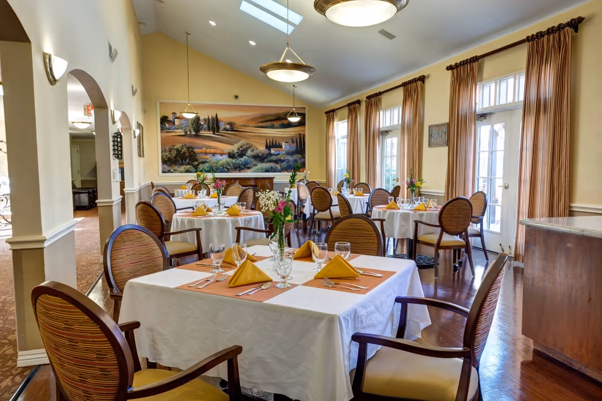 A bright and elegant dining room with several round tables covered in white tablecloths, each set with yellow napkins, glassware, and silverware. The room features wooden chairs with patterned upholstery, large windows with beige curtains, and a large landscape painting on the far wall. Ceiling lights and skylights provide ample lighting.