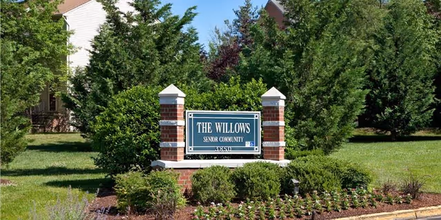 Brick entrance sign reading "The Willows Senior Community" surrounded by landscaped shrubs and trees.