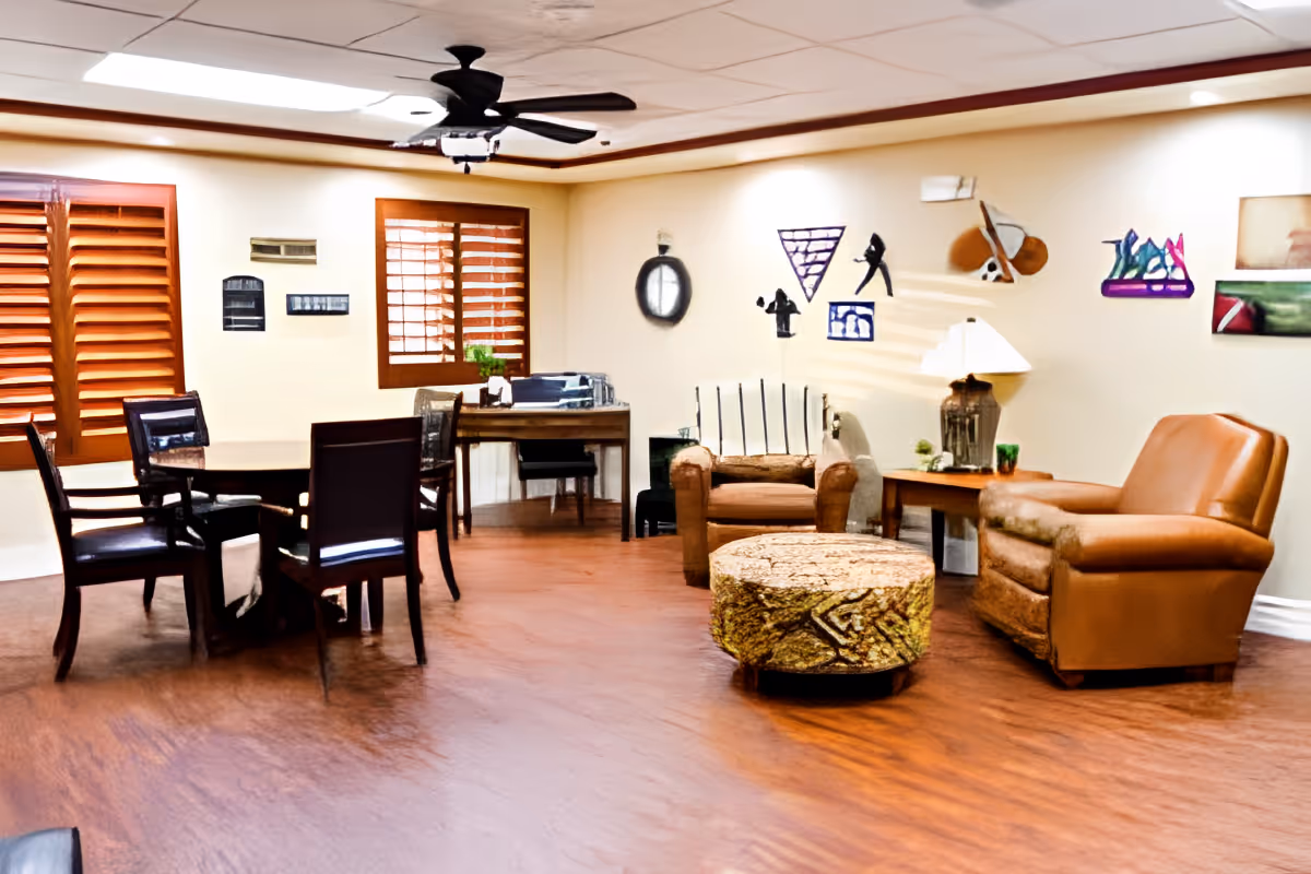A cozy living room area with wooden flooring, featuring a round table with four chairs on the left, two leather armchairs on the right, a patterned ottoman in the center, a small wooden desk with a chair near the back wall, and various decorative wall art. The room has wooden window shutters and a ceiling fan with lights.
