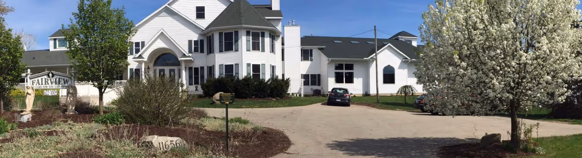 Exterior view of a large white multi-story building with multiple windows and a rounded turret-like section. The building is surrounded by a driveway, landscaped garden beds with bushes and trees, and a large tree with white blossoms on the right. A sign near the entrance reads 'Fairview Adult Foster Care'.