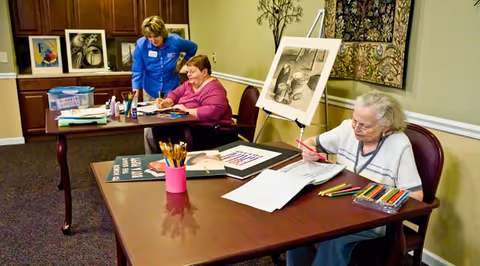Two elderly women seated at a table engaged in drawing and coloring activities with colored pencils and art supplies, while a staff member in a blue shirt stands nearby assisting. The room has framed artwork on the walls and a cabinet in the background.