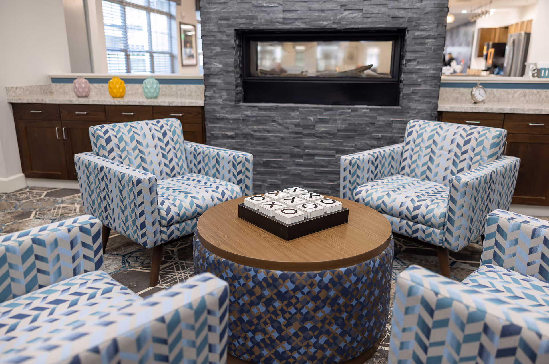 Contemporary seating area with four patterned armchairs arranged around a round wooden table topped with a tic-tac-toe game in front of a stone fireplace and cabinetry.