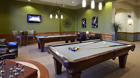 Interior view of a recreational room with two pool tables, a foosball table, a wall-mounted TV, bar stools, and comfortable seating. The walls are painted green with wood paneling, and there are modern pendant lights hanging from the ceiling.