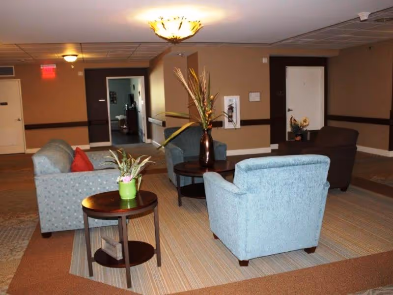 A seating area in a senior living facility with four upholstered chairs arranged around two small round tables. One table has a green potted plant, and the other has a tall vase with decorative dried plants. The room has beige walls, carpeted floors, and ceiling lights. Doors and a hallway are visible in the background.