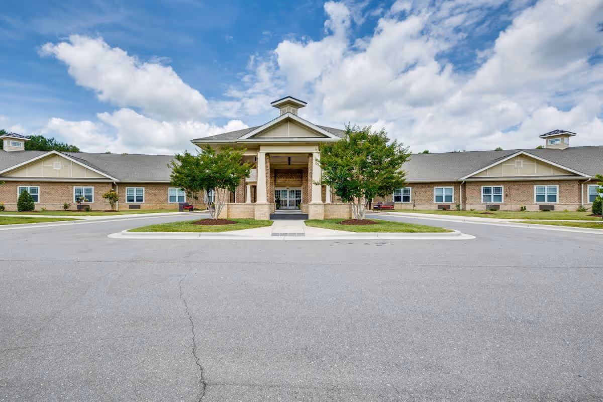Front entrance of a single-story brick assisted living facility with a porte-cochère, circular driveway, and landscaped trees under a partly cloudy sky.