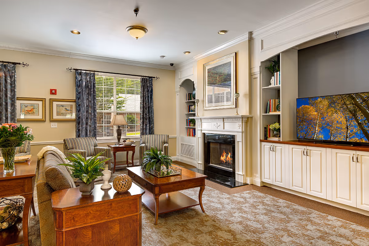 A cozy living room with a beige sofa, two striped armchairs, a wooden coffee table with plants, a fireplace with a framed picture above it, built-in white cabinets and shelves with books, a large flat-screen TV displaying a nature scene, and a window with blue patterned curtains letting in natural light.