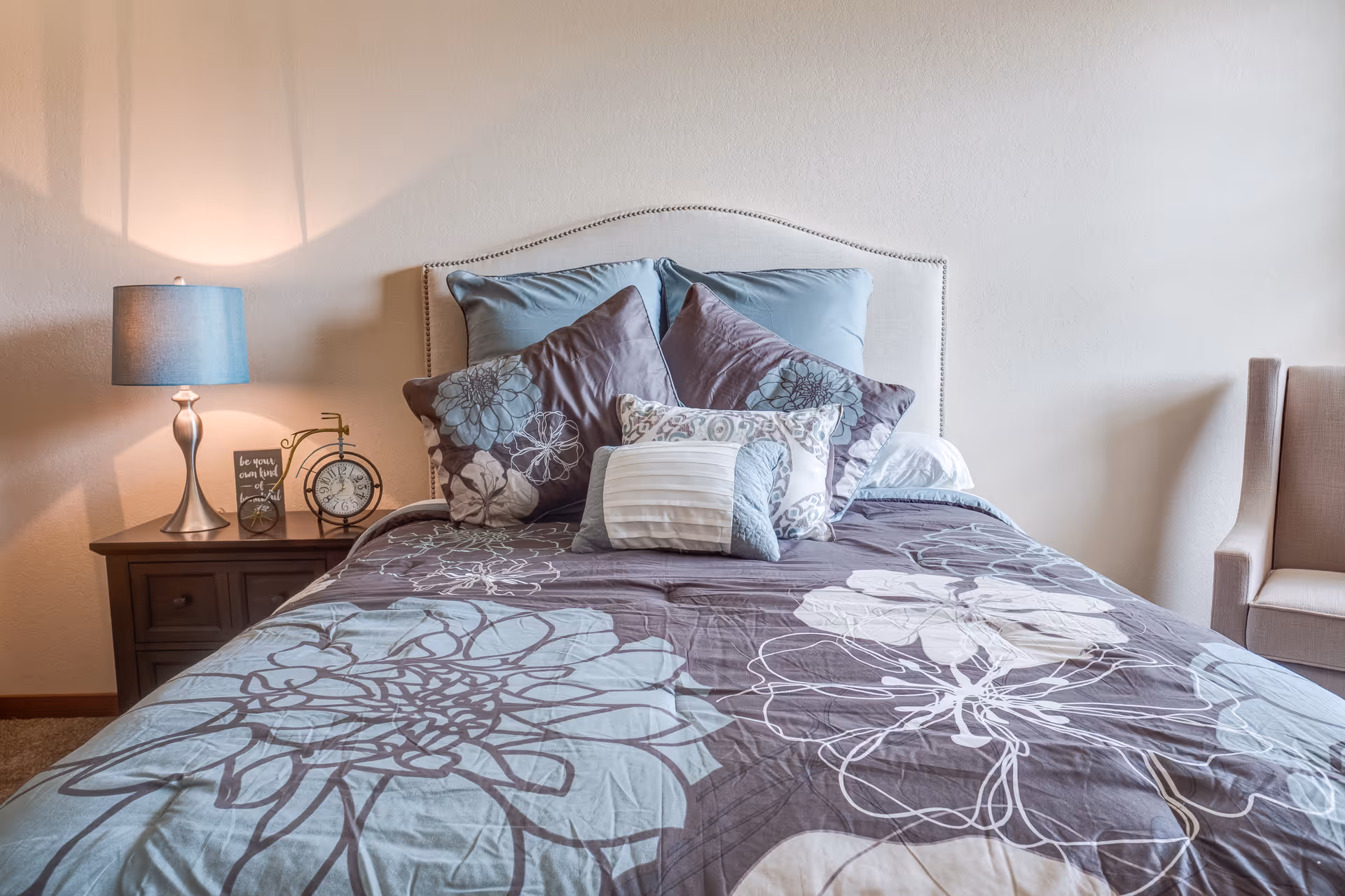 A neatly made bed with a floral patterned comforter and multiple pillows in shades of blue and gray. To the left of the bed is a wooden nightstand with a blue lamp, a small decorative clock, and a framed inspirational quote. To the right of the bed is a beige upholstered armchair against a plain light-colored wall.