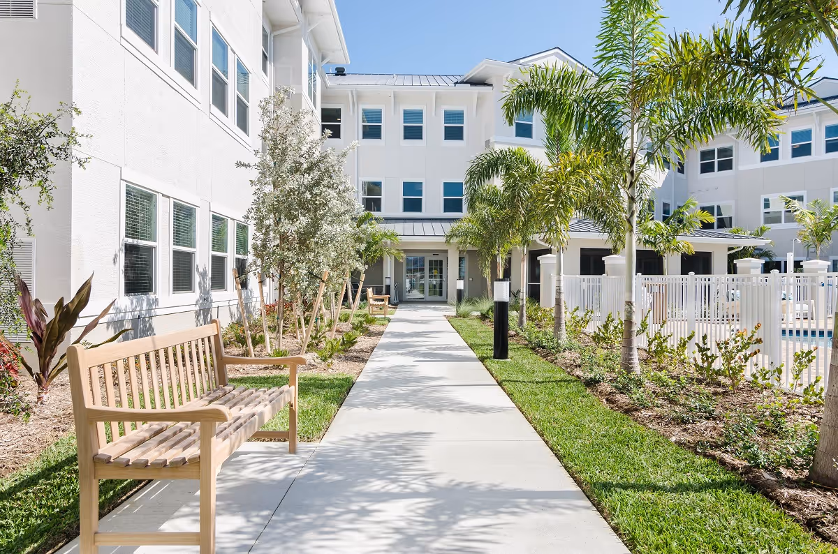 Outdoor walkway at American House Fort Myers with wooden benches along the path, palm trees, and a white building with multiple windows under a clear blue sky.