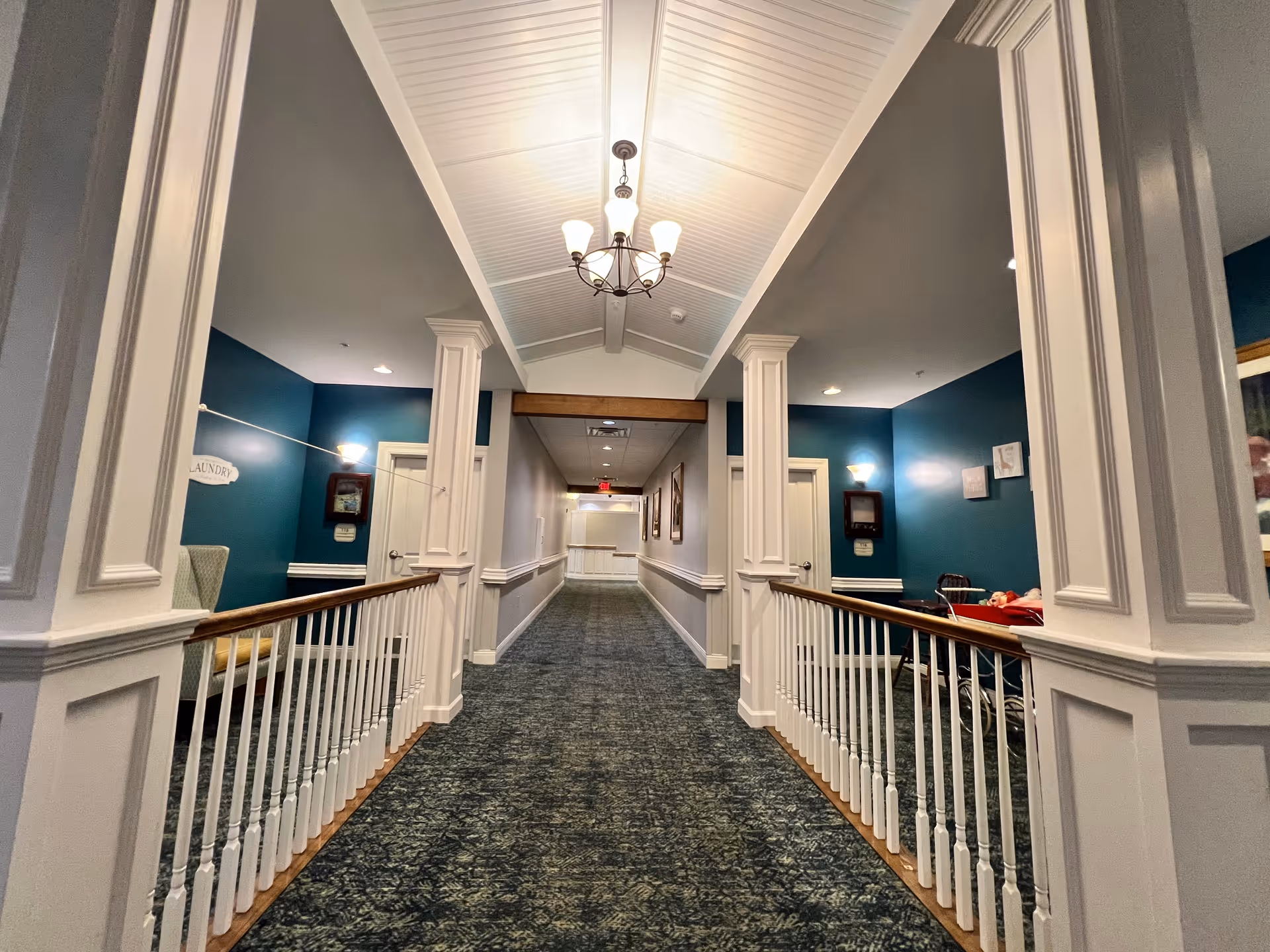 A well-lit hallway in a senior living facility with teal walls, white trim, and carpeted floor. The hallway features white railings on both sides, decorative columns, and a chandelier hanging from a vaulted ceiling. There are doors and framed pictures along the walls, and a sign that reads 'Laundry' is visible on the left side.