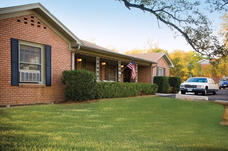 Red brick senior living facility exterior with front porch, flag, hedges, and parked cars.