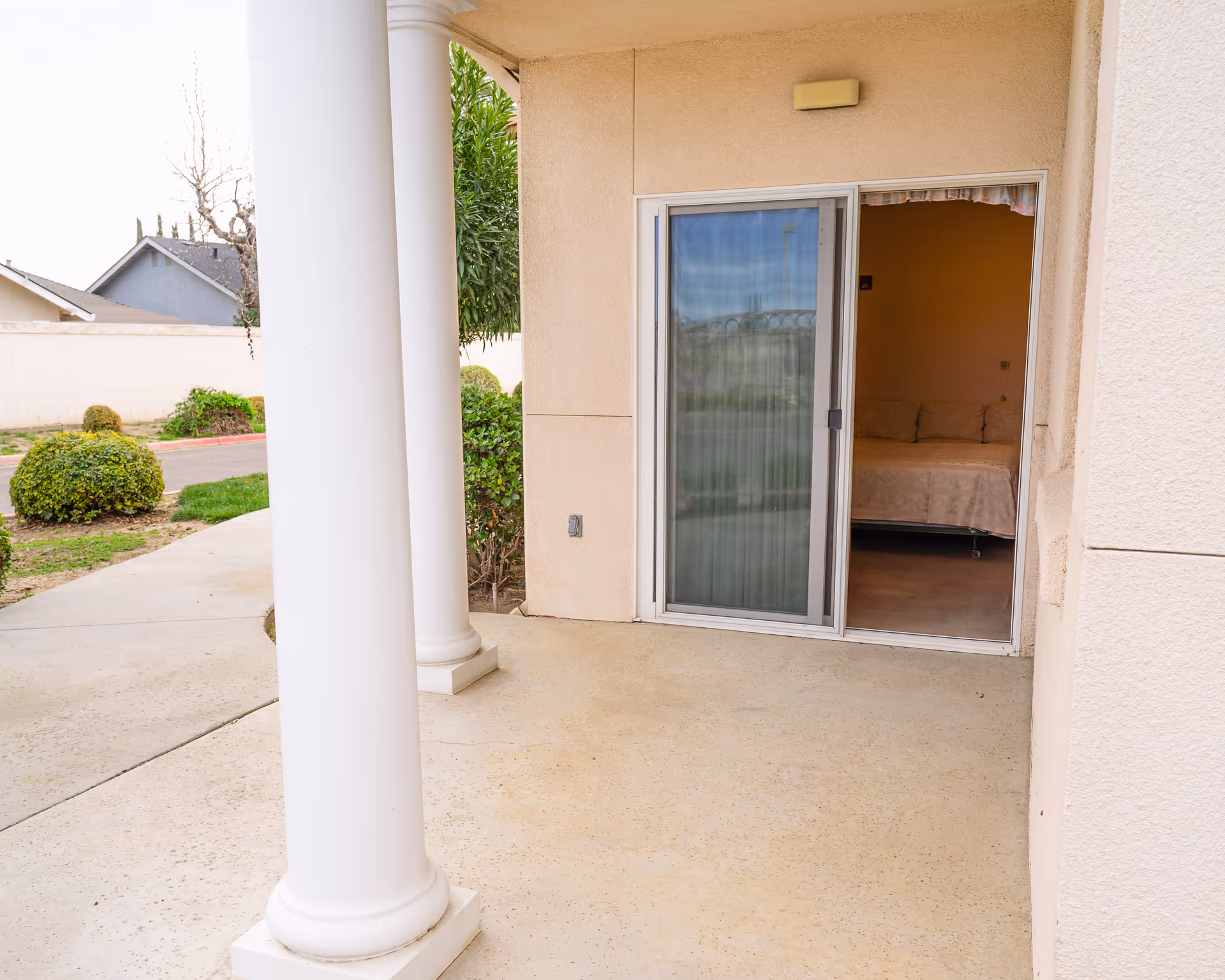 Outdoor patio area with two white columns and a sliding glass door leading into a room with a bed visible inside. There are bushes and a sidewalk outside, along with a neighboring house and a white wall in the background.