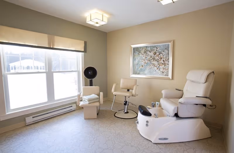 A bright room with beige walls and a large window covered by a beige valance. The room contains a white pedicure spa chair with foot bath, a salon chair, a small chair with folded towels, and a hair dryer hood. A framed floral painting hangs on the wall.