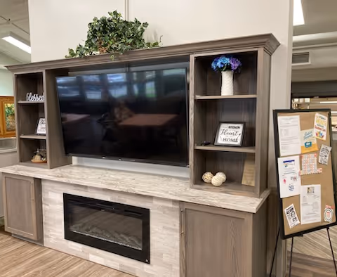 A modern entertainment center with a large flat-screen TV mounted in the middle, surrounded by shelves holding decorative items including a plant, a vase with purple flowers, and small ornaments. Below the TV is an electric fireplace. To the right of the entertainment center is a bulletin board with various notices and papers pinned on it.