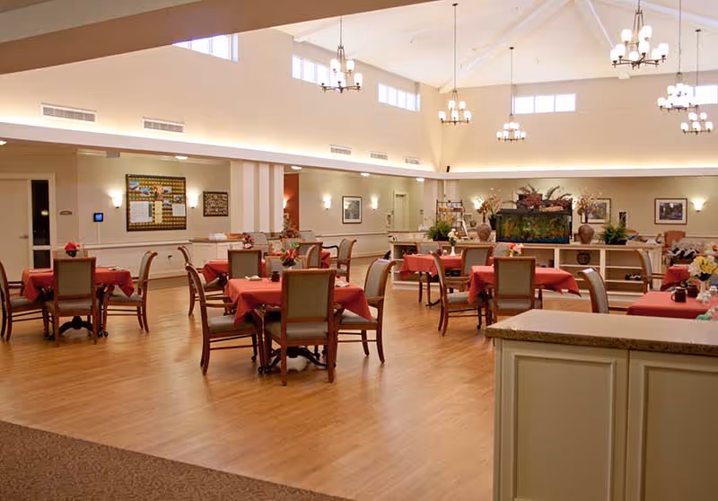 A spacious dining room with multiple tables covered in red tablecloths, each surrounded by wooden chairs with cushioned seats. The room has high ceilings with several chandeliers, large windows near the ceiling allowing natural light, and light-colored walls adorned with framed pictures. There is a large fish tank on a shelf against the far wall, and the floor is made of light wood.