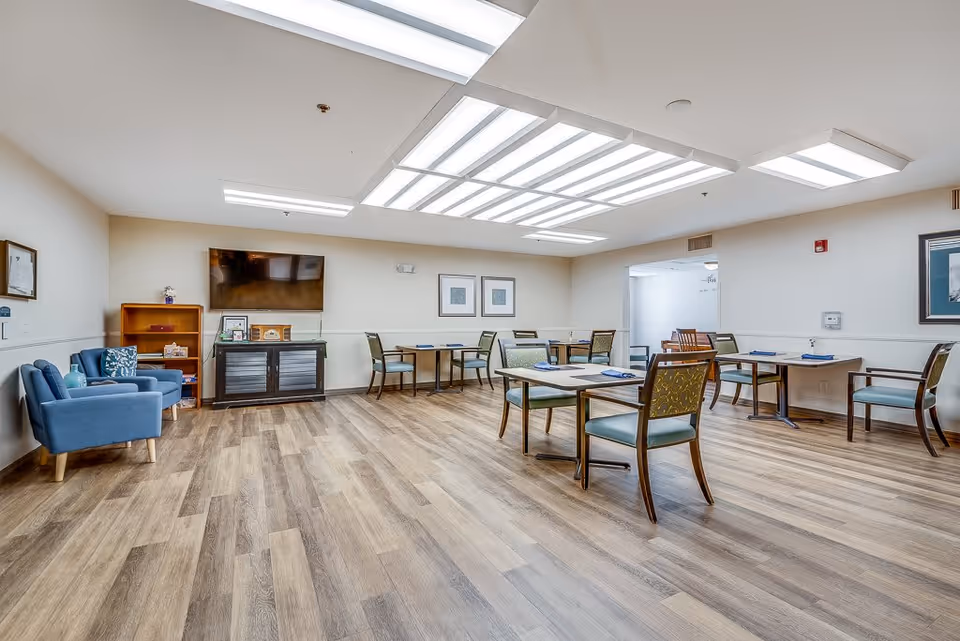 A spacious, well-lit dining room with several tables and chairs arranged neatly. The room features wood flooring, a large flat-screen TV mounted on the wall, and two blue armchairs with a small wooden shelf nearby. The ceiling has multiple rectangular fluorescent light panels, and the walls are decorated with framed pictures.