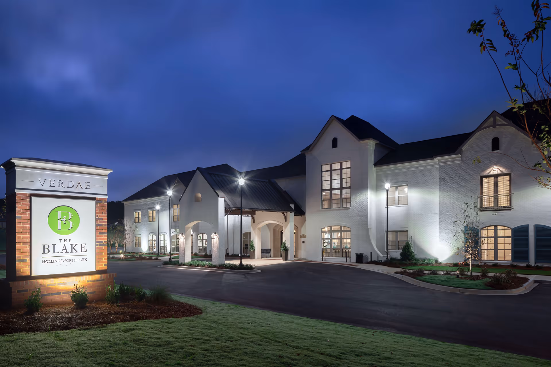 Exterior view of The Blake at Hollingsworth Park senior living facility at dusk, showing a large white building with multiple windows and a covered entrance, illuminated by outdoor lights. A sign with the facility's name is visible on the left side of the image.