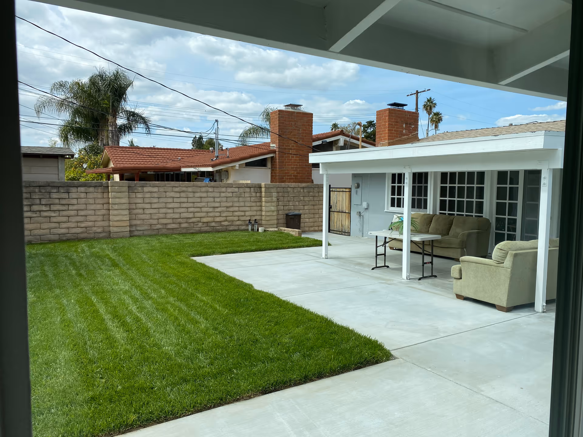 View of a backyard with a well-maintained green lawn, a concrete patio area with a covered seating space that includes a sofa, an armchair, and a table. The yard is enclosed by a brick wall, and neighboring houses with red-tiled roofs and palm trees are visible in the background under a partly cloudy sky.