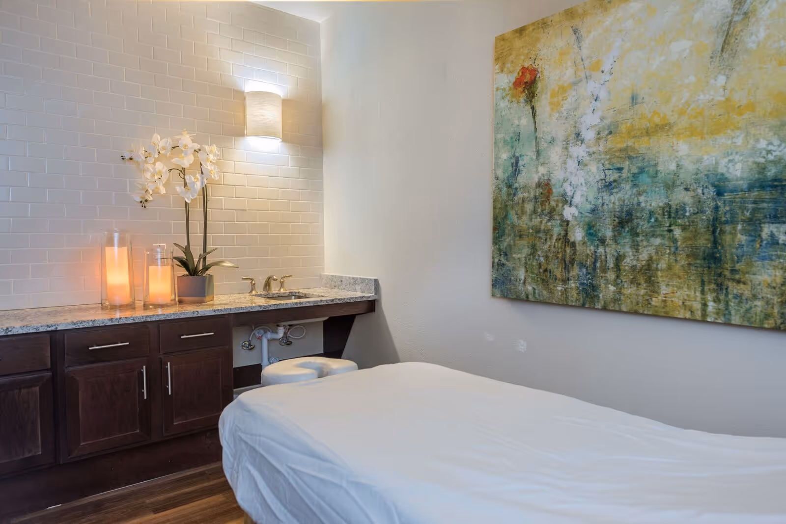 A serene treatment room with a massage table covered in a white sheet, a countertop with a sink, two lit candles, and a white orchid plant. The wall behind the countertop is tiled with white subway tiles, and a modern wall light fixture is mounted above. A large abstract painting with green, yellow, and red tones hangs on the adjacent wall.