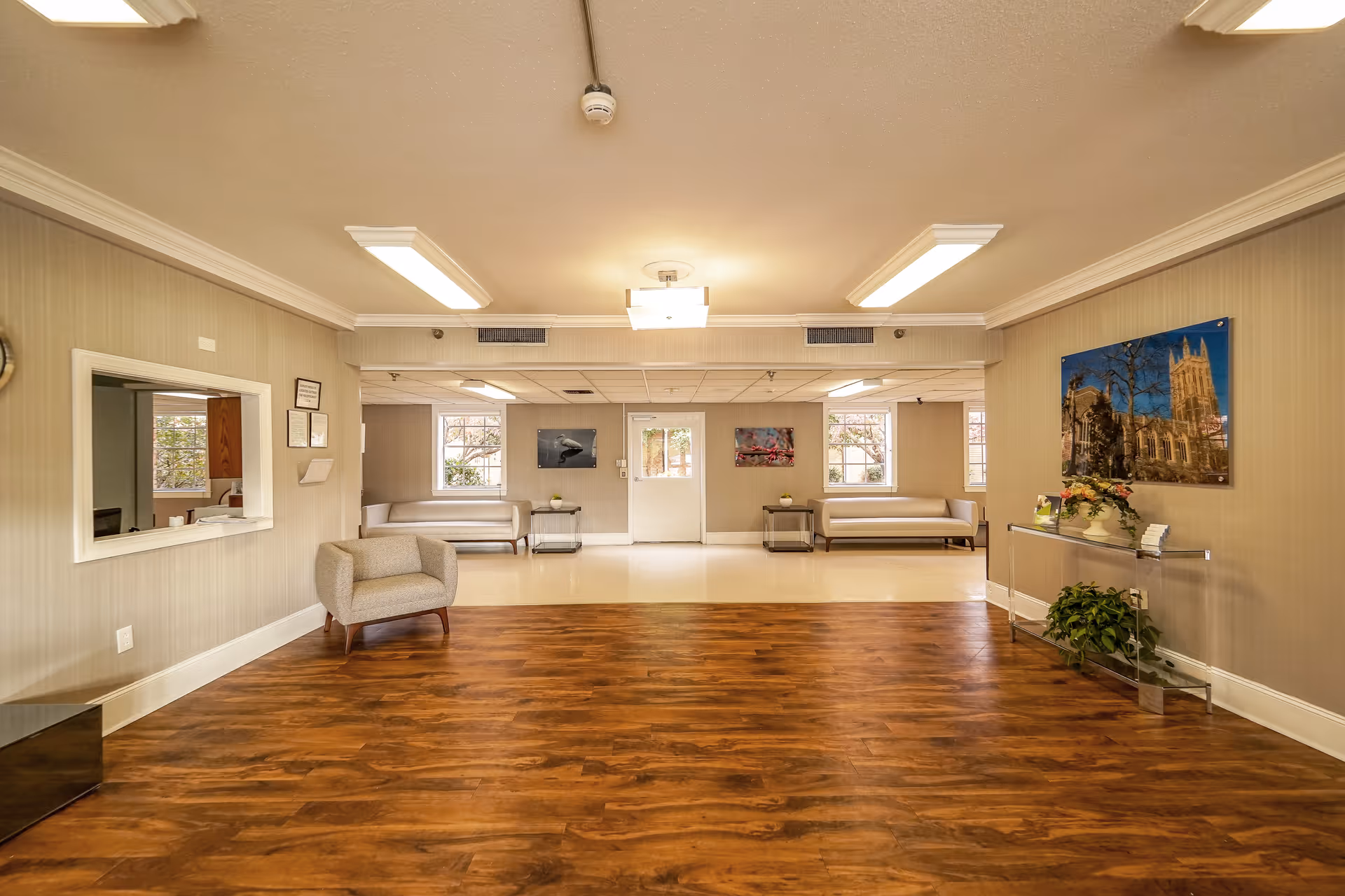 A spacious and well-lit common area in a senior living facility with wooden flooring in the foreground and a tiled floor in the background. The room features beige walls, two gray sofas along the back wall, a single armchair on the left, a glass console table with flowers and a plant on the right, and framed artwork on the walls. There is a window-like opening on the left wall and a door in the center back wall.