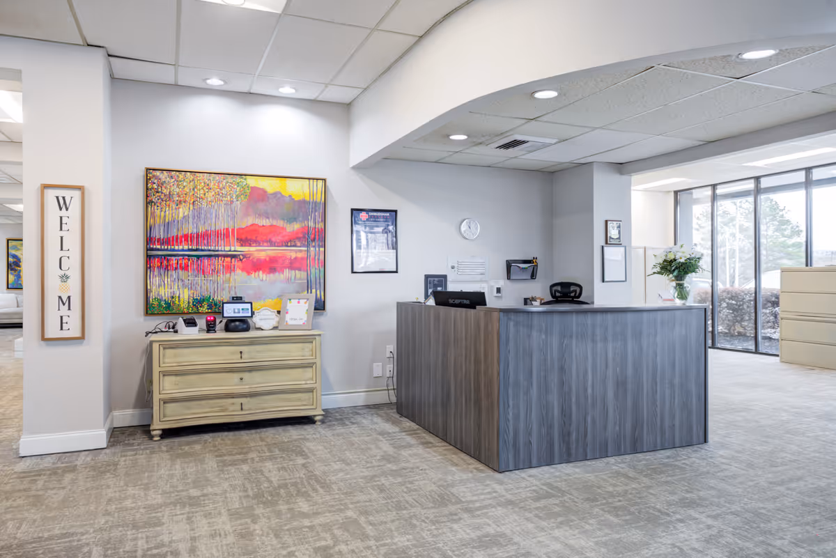 Reception area in Commonwealth Senior Living At Oak Ridge featuring a wooden reception desk with a chair behind it, a small clock on the wall, a colorful landscape painting above a three-drawer cabinet, and a vertical welcome sign on the wall. Large windows allow natural light into the space.