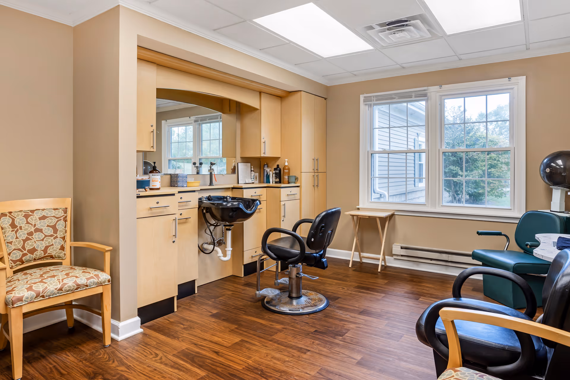 Interior view of a hair salon area in a senior living facility with a black salon chair in front of a black wash basin, light wood cabinetry with a large mirror, and a window showing the outside. There are additional chairs with patterned upholstery and a green salon chair with a hair dryer nearby. The floor is wood and the walls are beige.